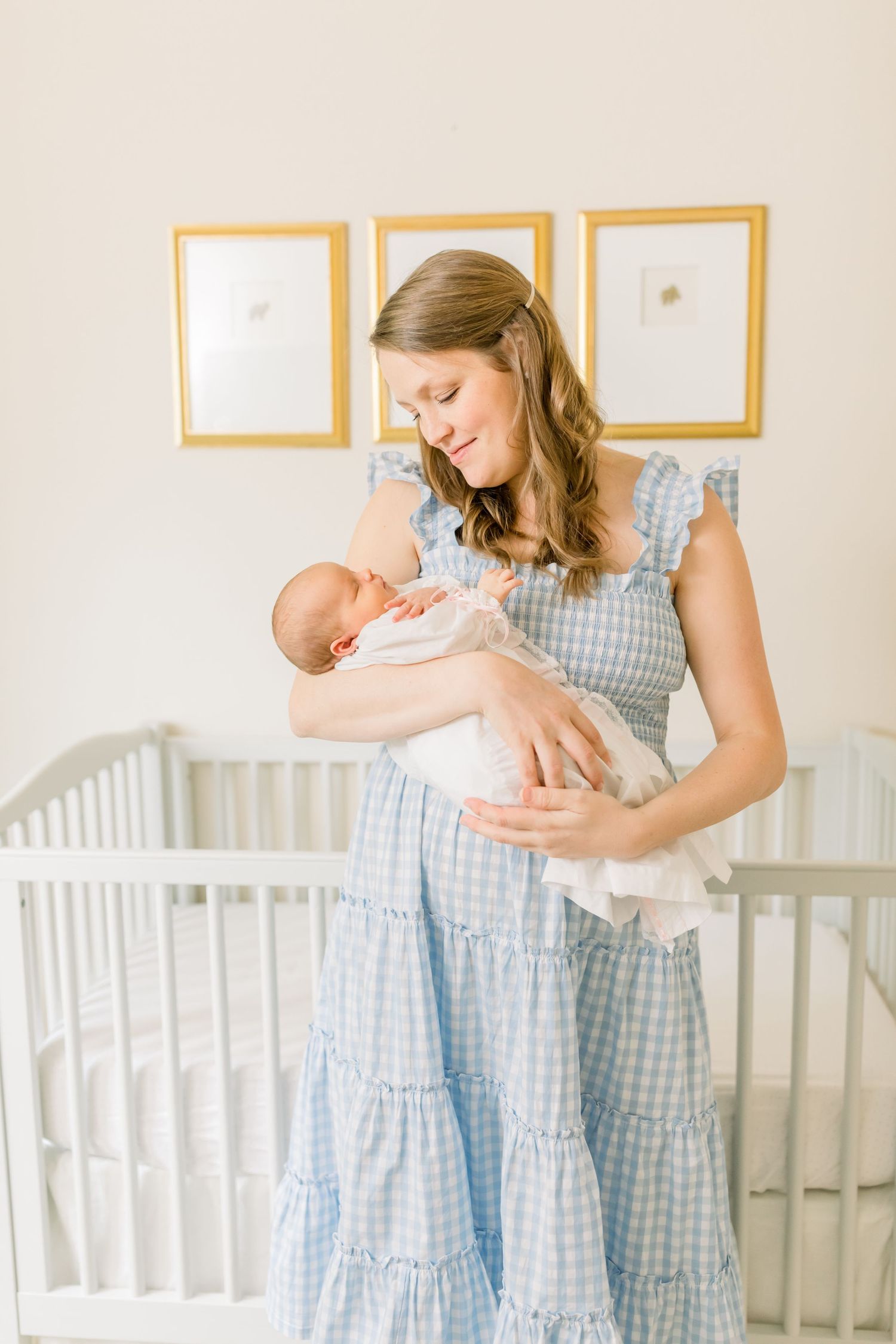 A figure in a blue gingham dress gazes lovingly at a newborn baby in a bright nursery setting.