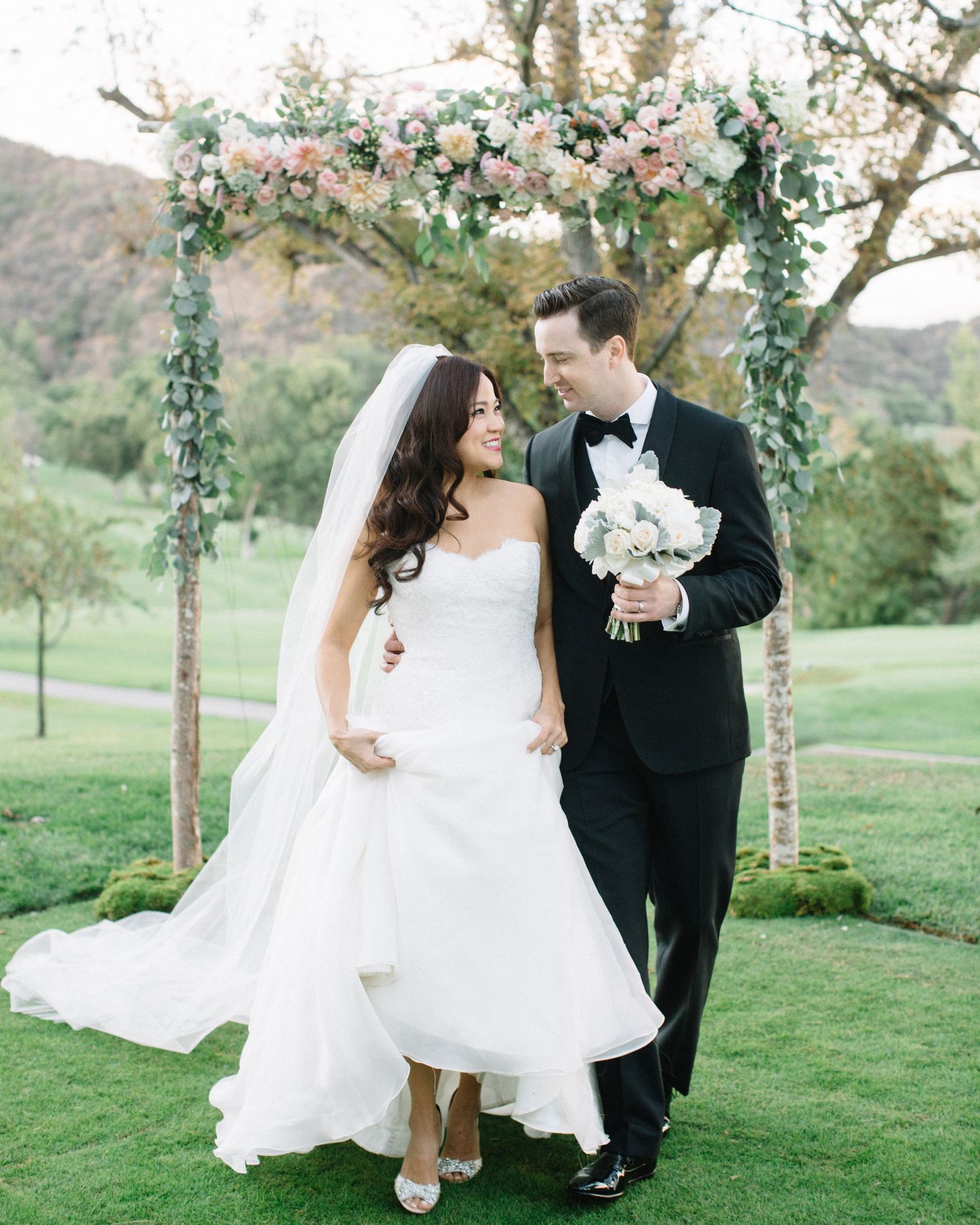 Romantic outdoor wedding portrait of a smiling bride and groom walking on a lawn past a lush floral arch.