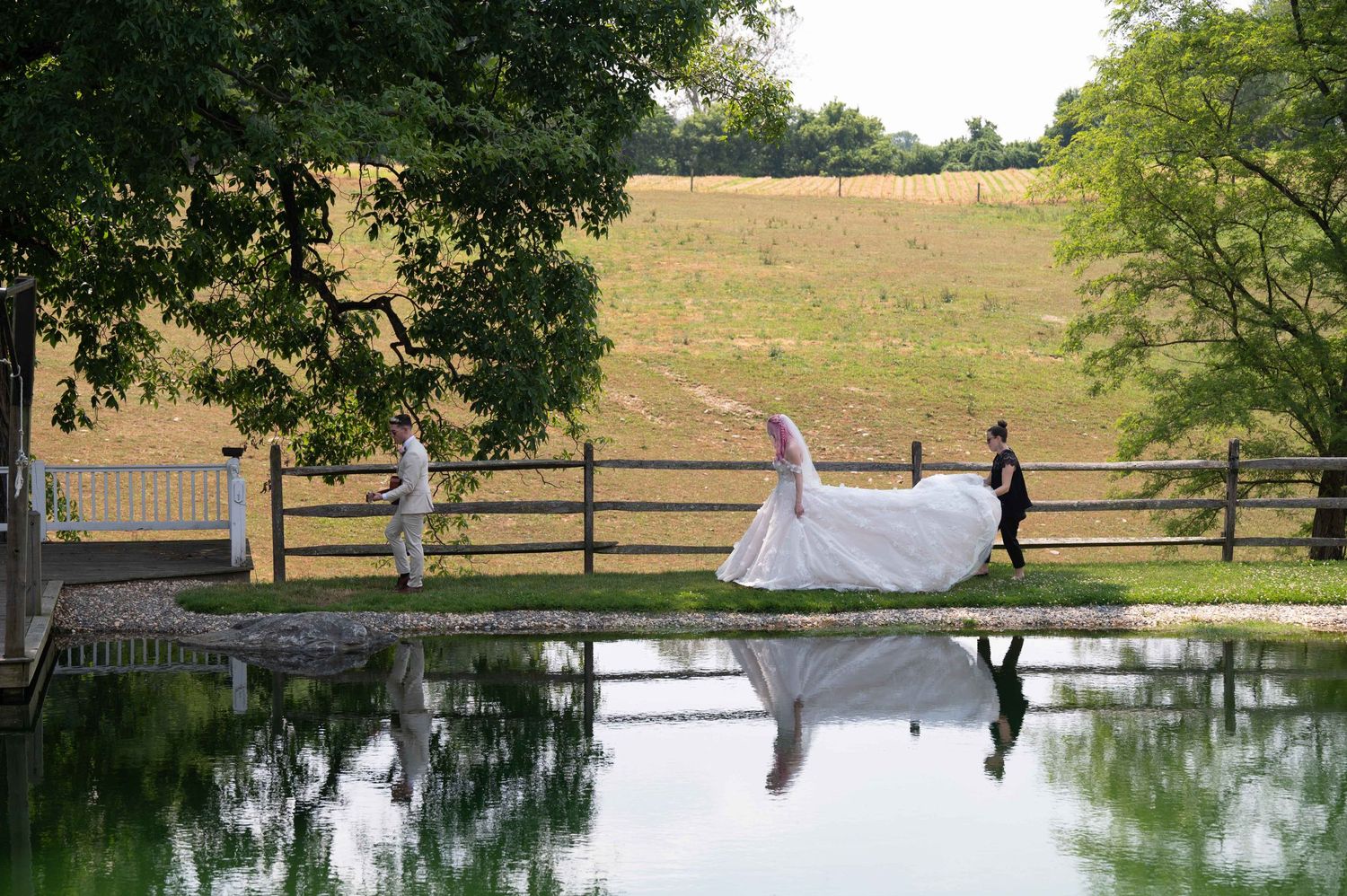 Wedding scene reflected in still pond water with wooden fence and countryside in background.