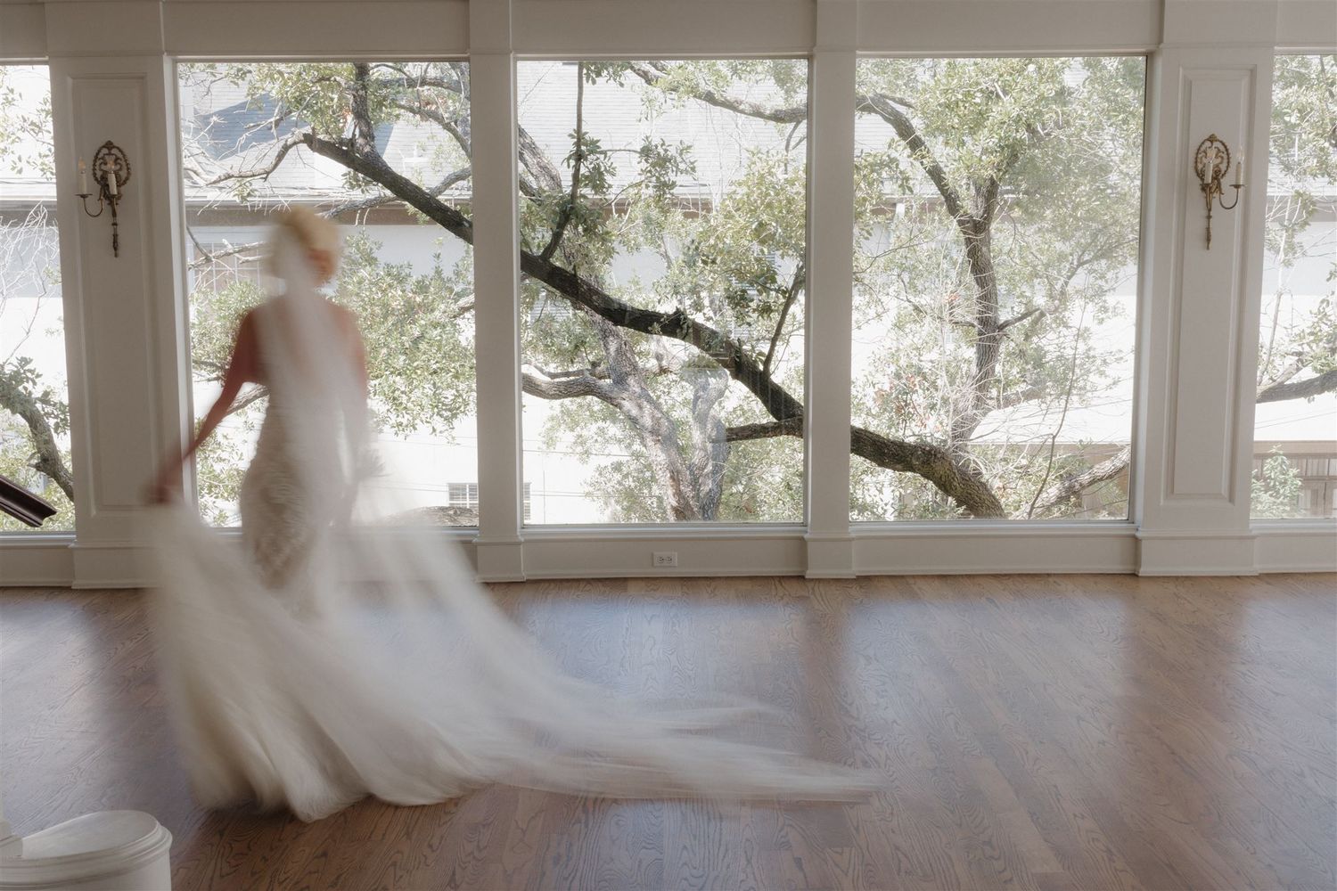Ethereal silhouette of a bride in motion against large windows with trees visible outside.