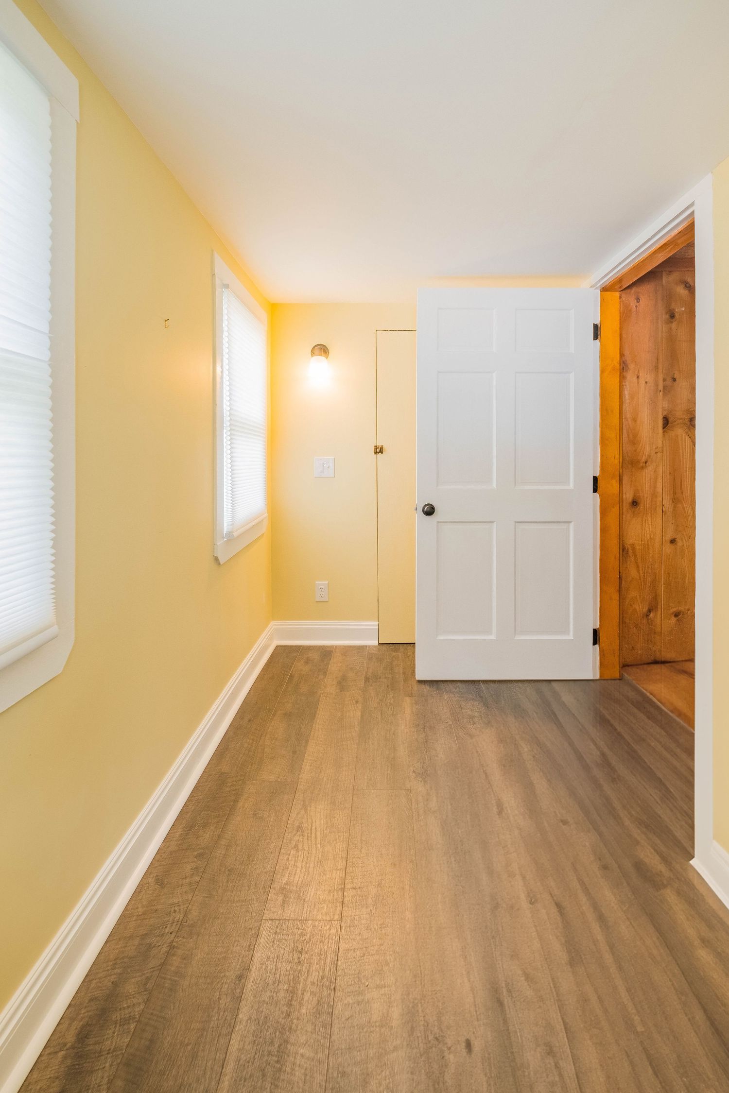Empty hallway with yellow walls, white trim, and modern wood-look flooring leading to white and wooden doors.