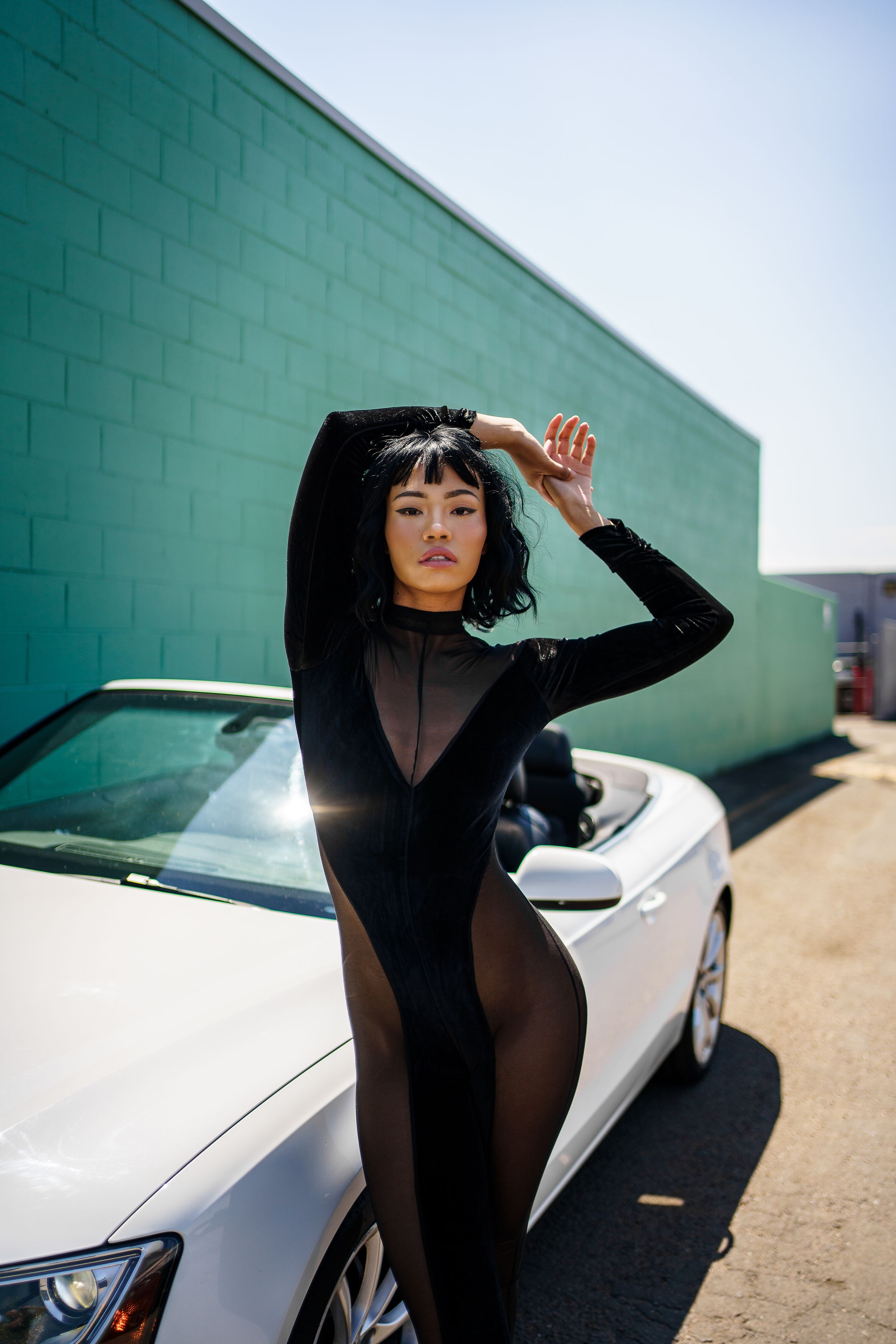 Fashion model in black sheer outfit poses against turquoise wall with white car in background.