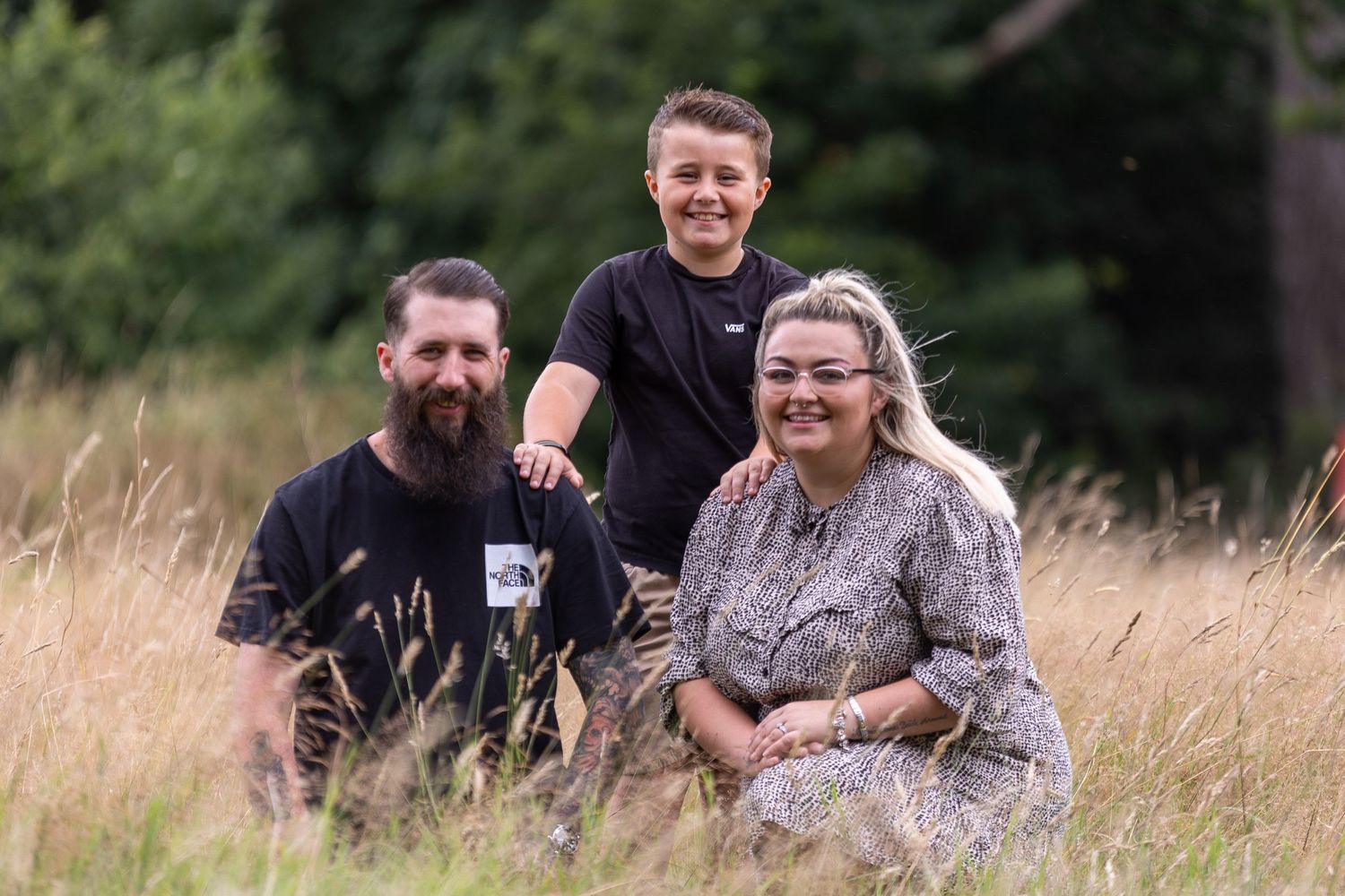 Three people pose together in a sunny field with tall grass and trees.