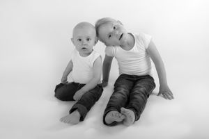 Deux enfants en débardeurs blancs et pantalons noirs, assis ensemble sur un fond blanc, dans une photographie en noir et blanc.