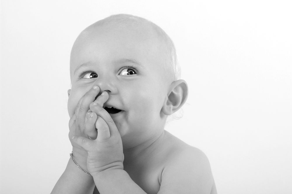 Photo en noir et blanc d’un bébé mignon regardant vers le haut, les mains près de la bouche, avec une expression joueuse.