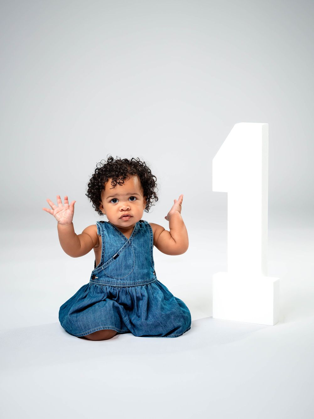 Baby in denim dress sits next to large white number one against gray background.