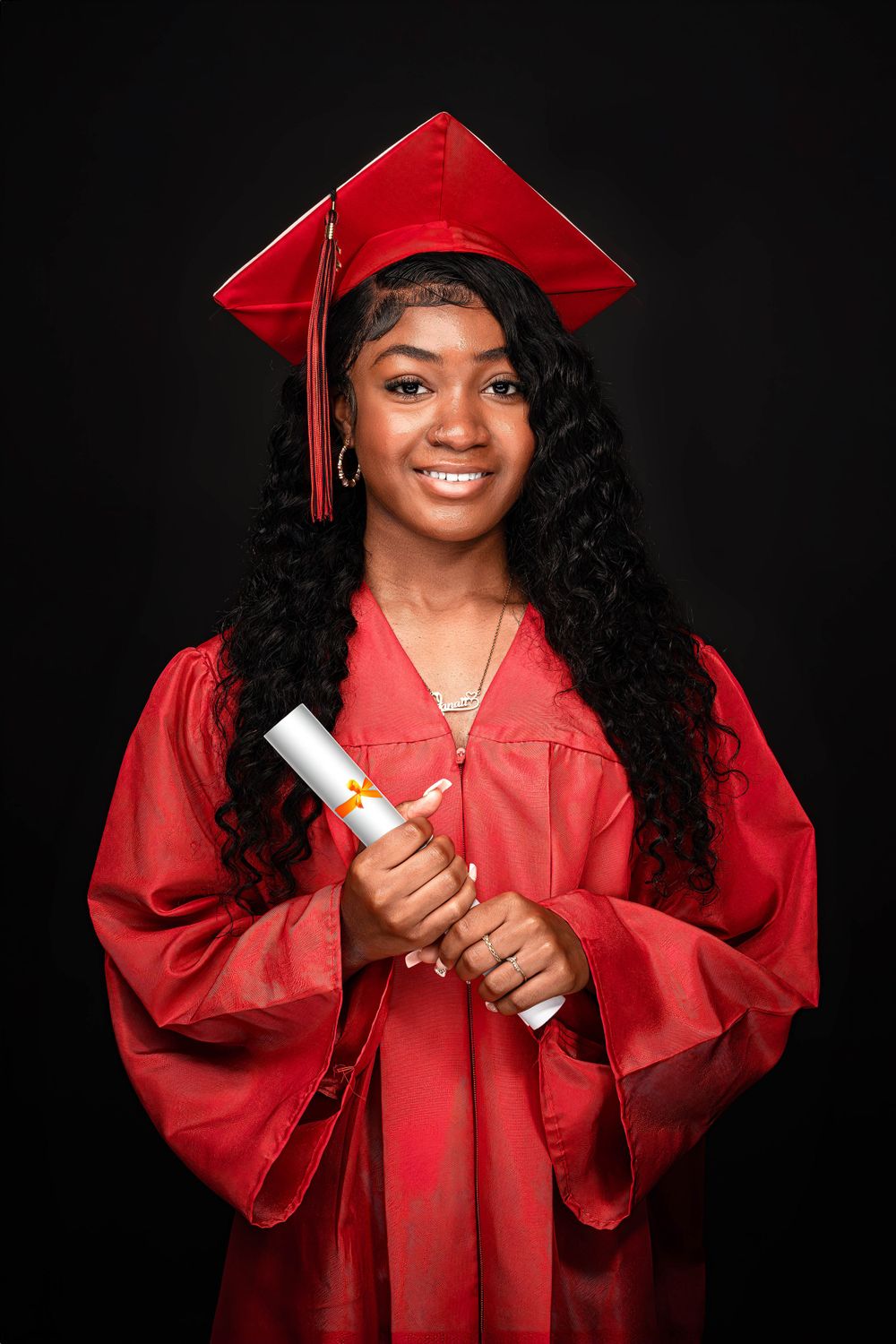 Graduate wearing red cap and gown holds diploma scroll while smiling against dark background.