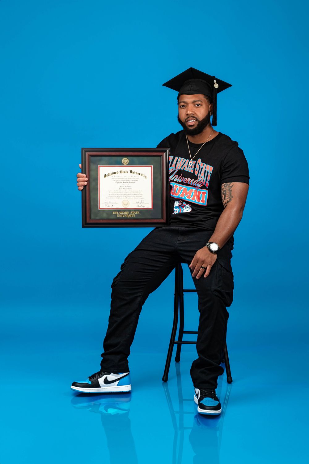 A person sits on a black stool wearing a black outfit and graduation cap while displaying a framed certificate against a blue backdrop.