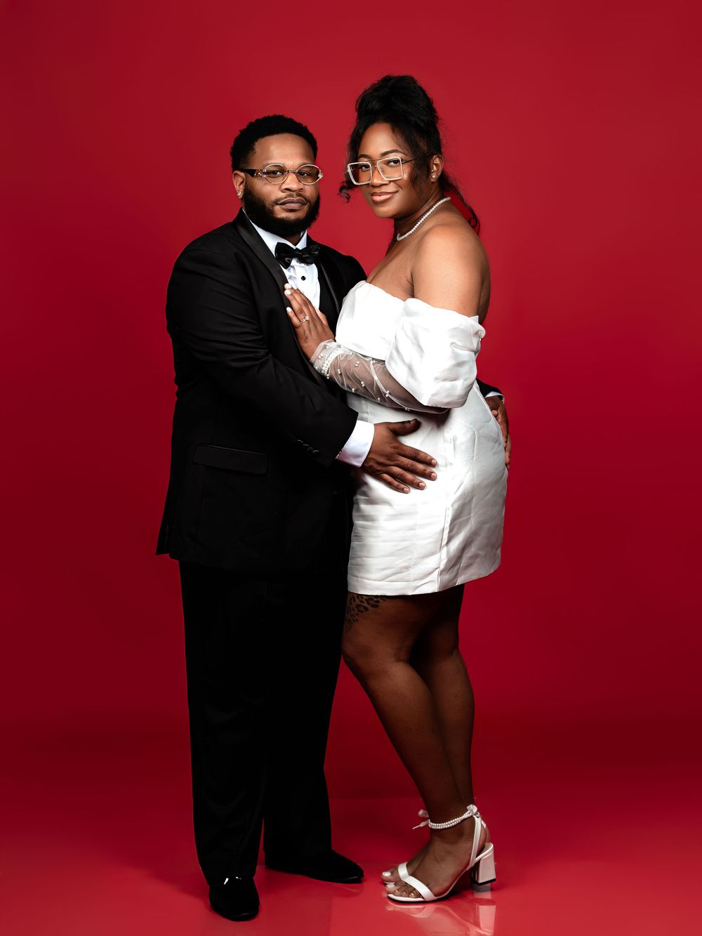 A couple poses together against a red backdrop in a romantic photoshoot series.