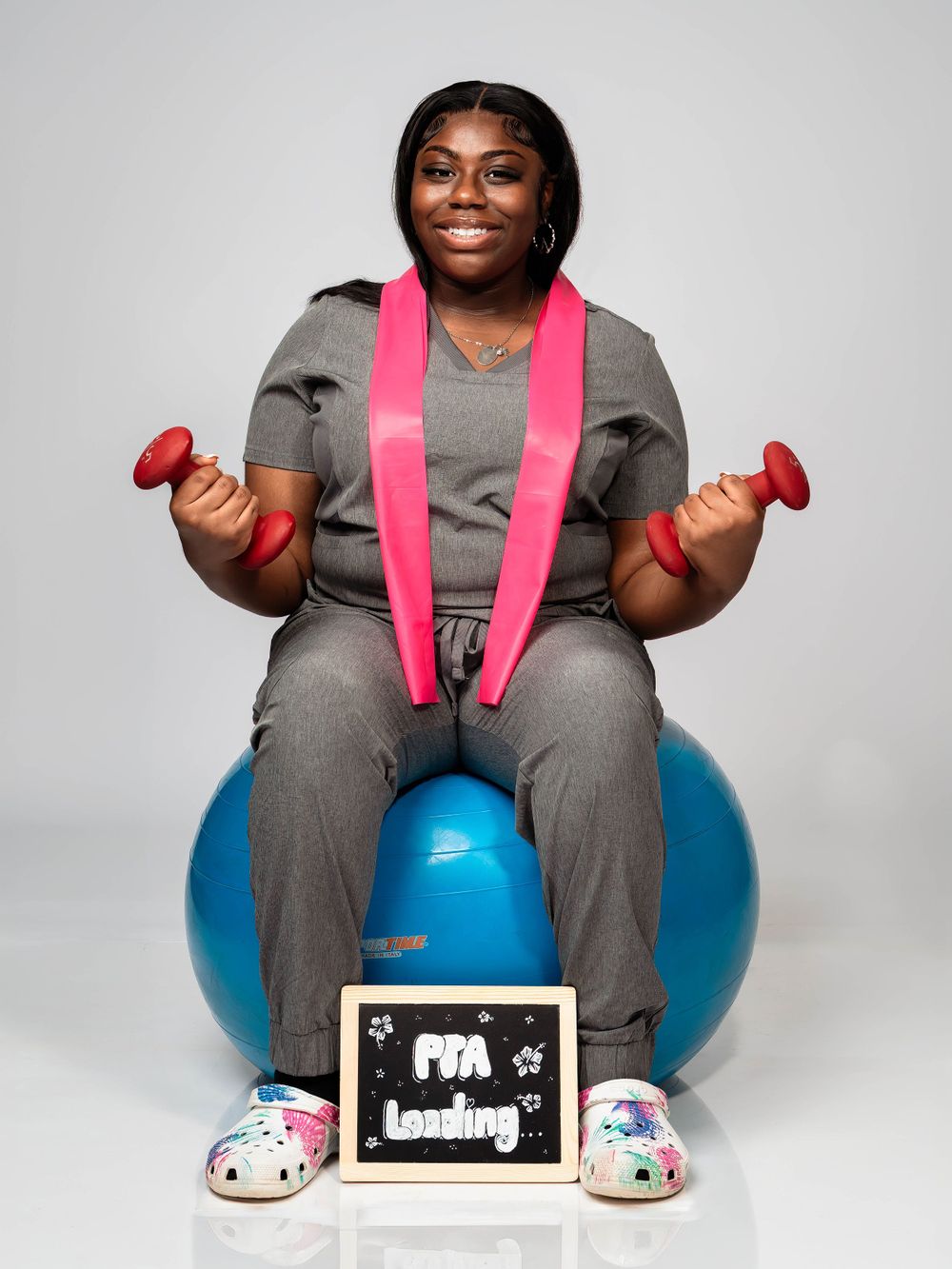 Physical therapist sits on exercise ball holding dumbbells in workout attire.