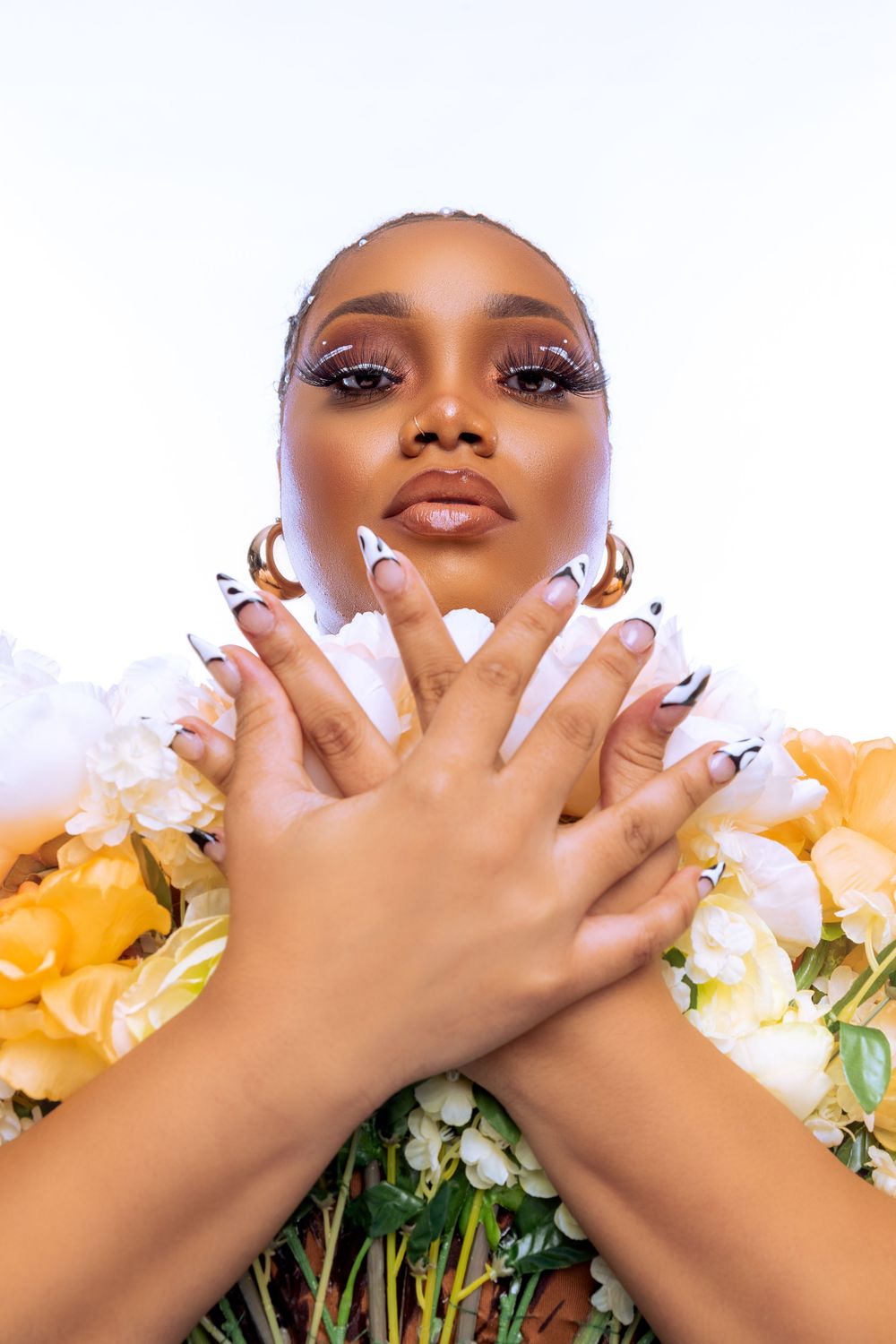 Black woman with dramatic lashes and long nails poses surrounded by yellow and white flowers.