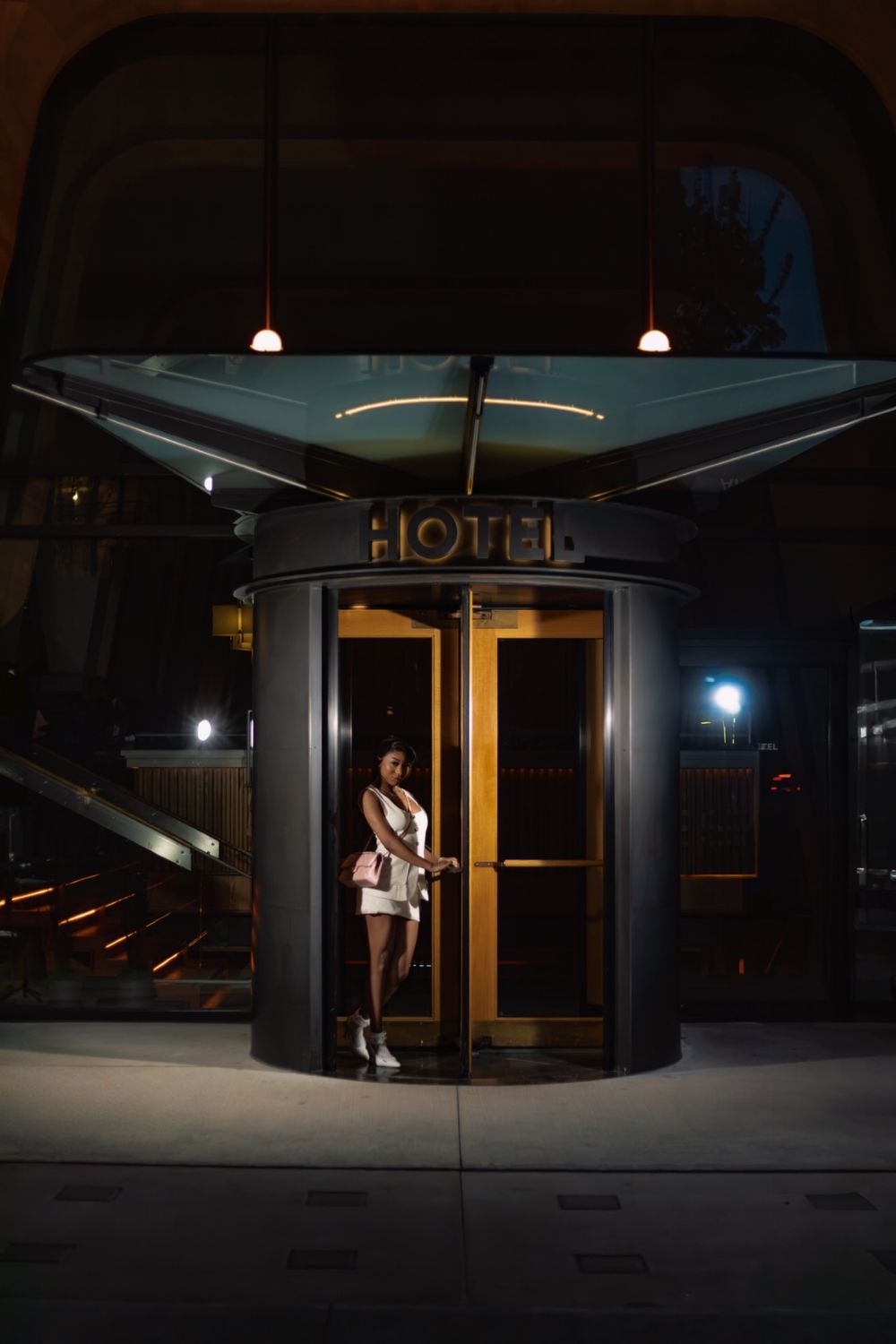 Woman in white dress exiting a luxury hotel through revolving gold doors at night.