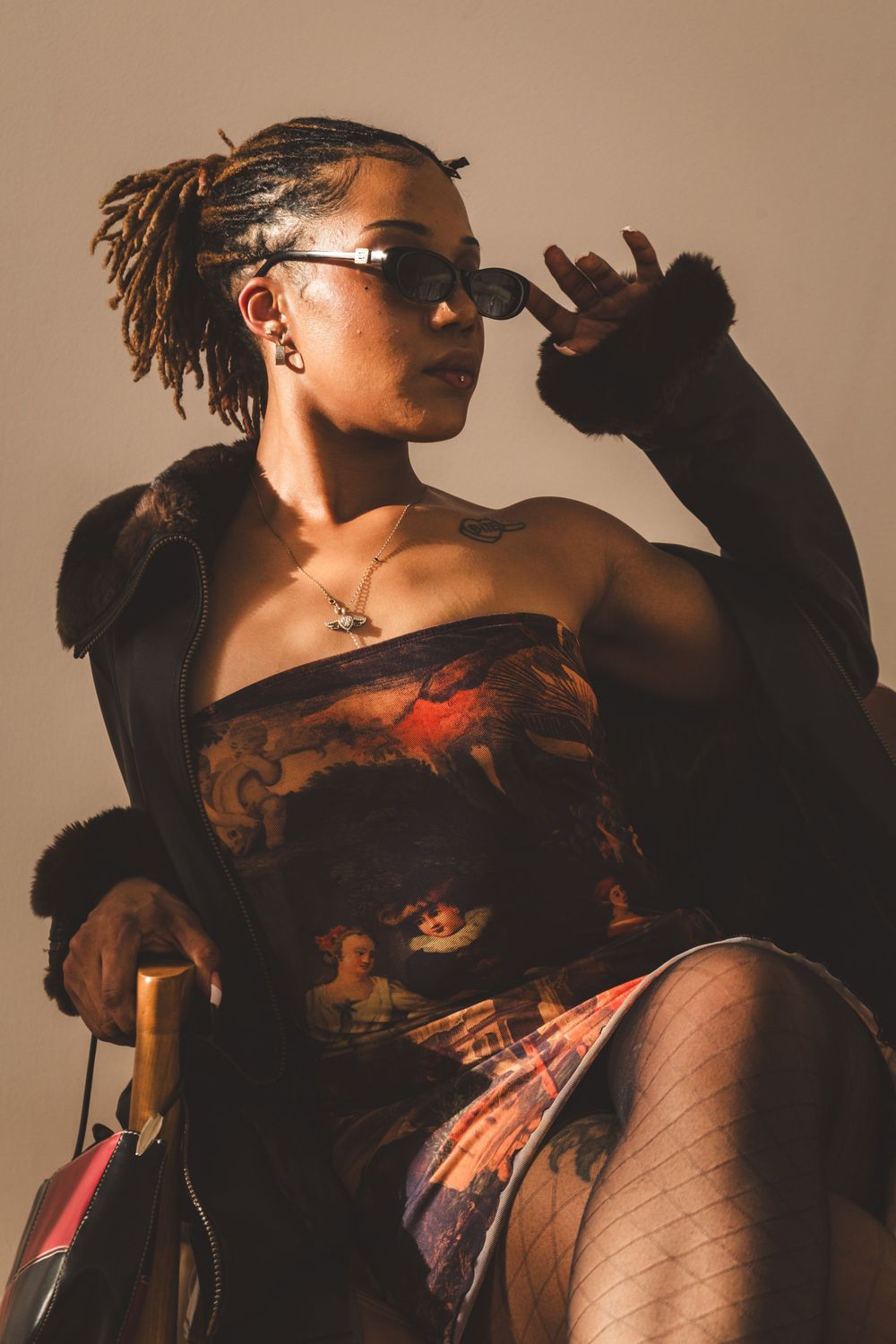 Stylish woman in printed strapless dress, fur coat, and sunglasses seated confidently in warm studio lighting.
