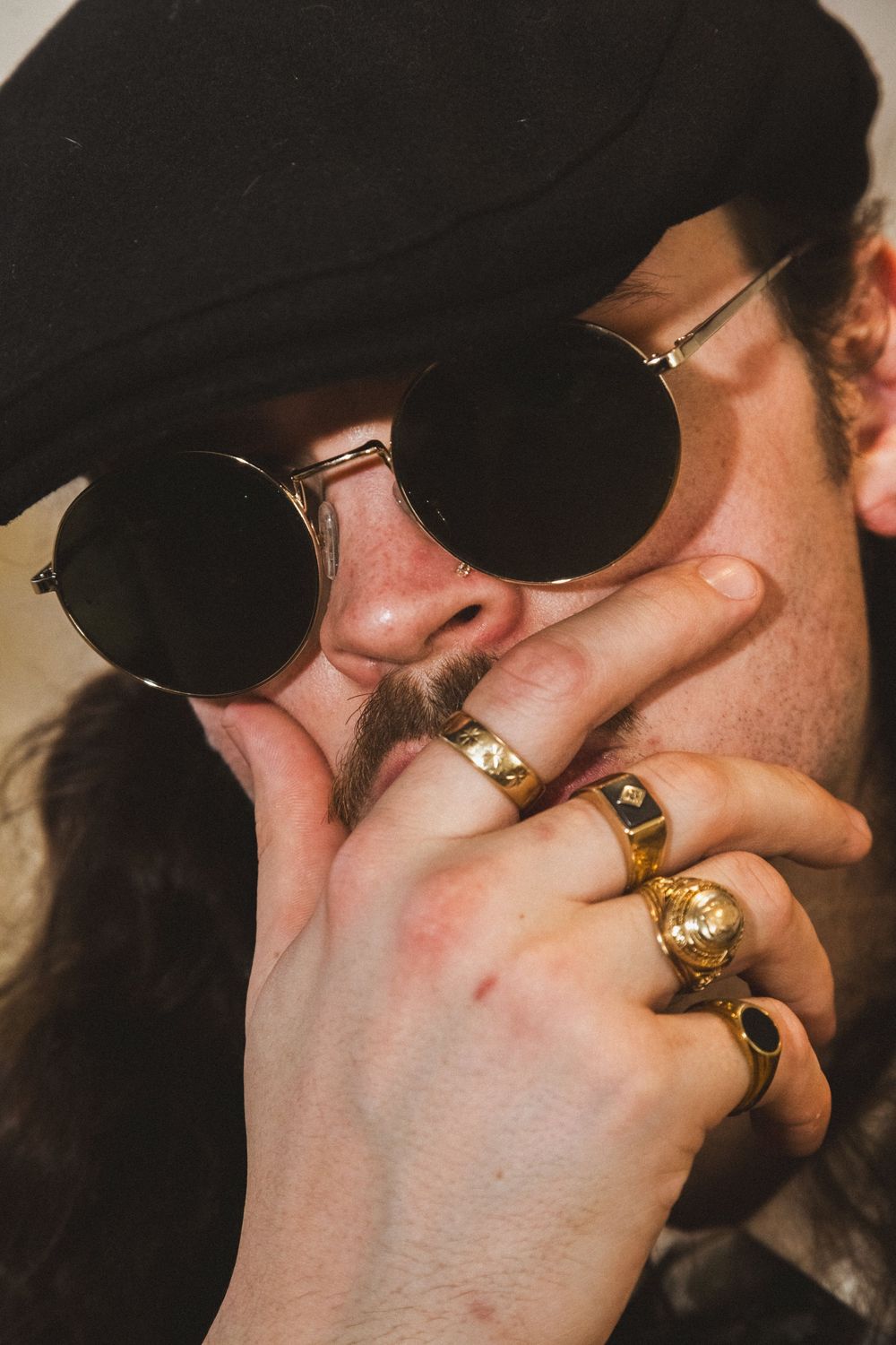 Man with round black sunglasses, black hat, soul patch, and multiple gold rings poses mysteriously.