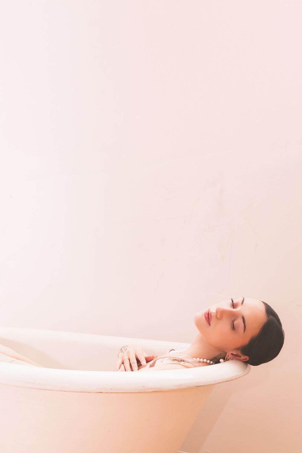 Woman relaxing in a vintage bathtub wearing pearl jewelry with eyes closed in a soft pink serene setting.