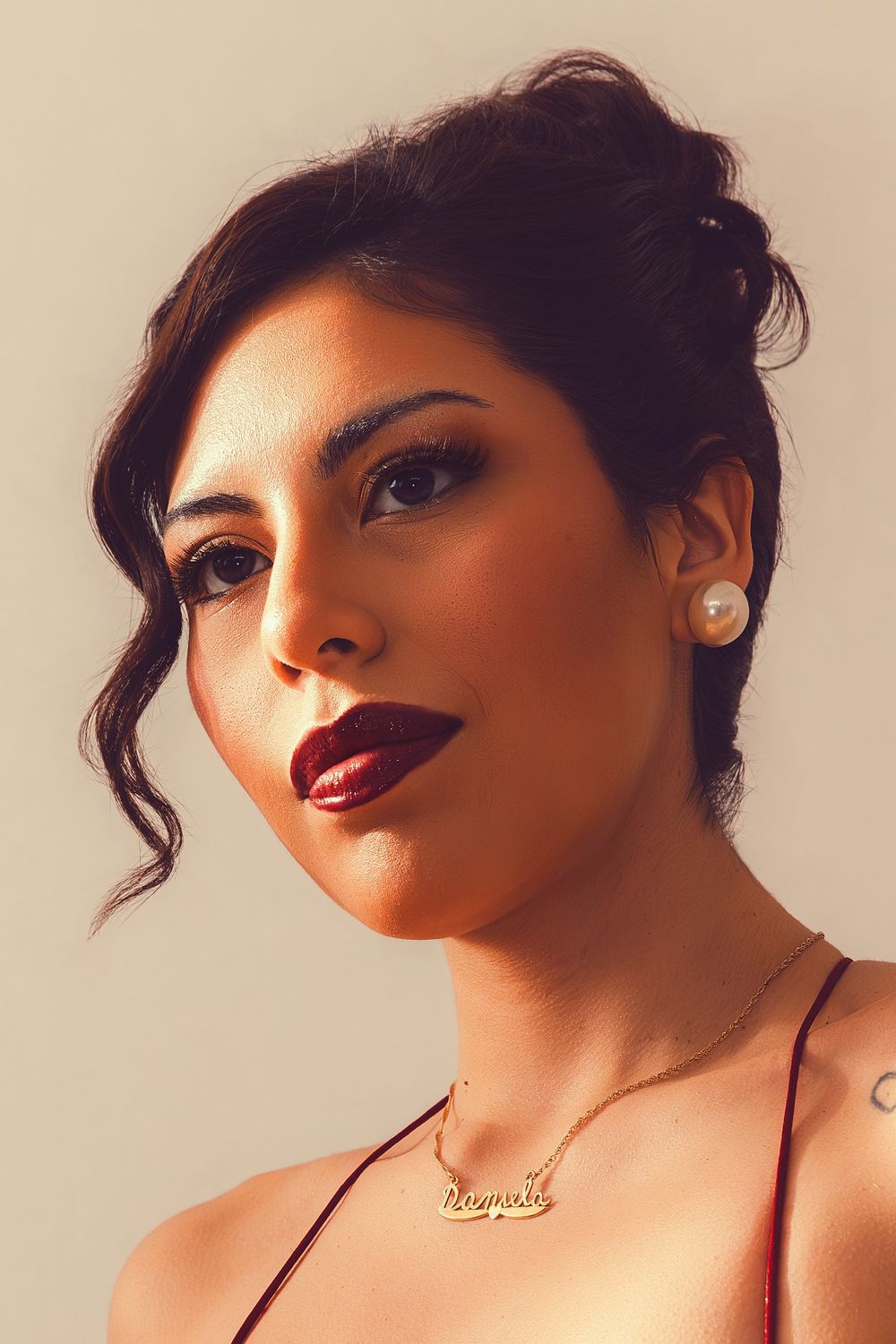 Woman with dark updo hair, pearl earrings, gold name necklace, and deep red lips poses against neutral background.