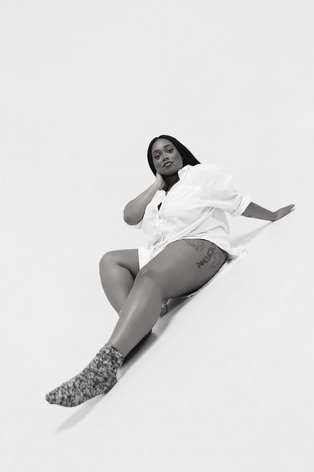 Black and white photo of a woman in a white shirt and socks sitting on a white floor in a relaxed pose.