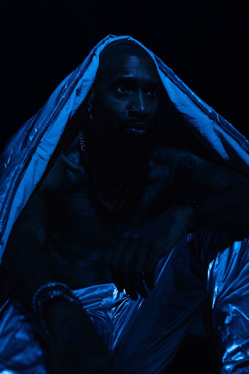 Man wrapped in silver plastic sheet under dramatic blue lighting in dark studio setting.