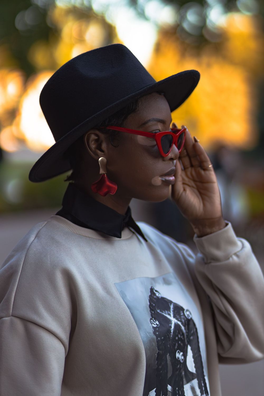 Stylish Black woman in beige sweatshirt wears black fedora and red cat-eye sunglasses outdoors.