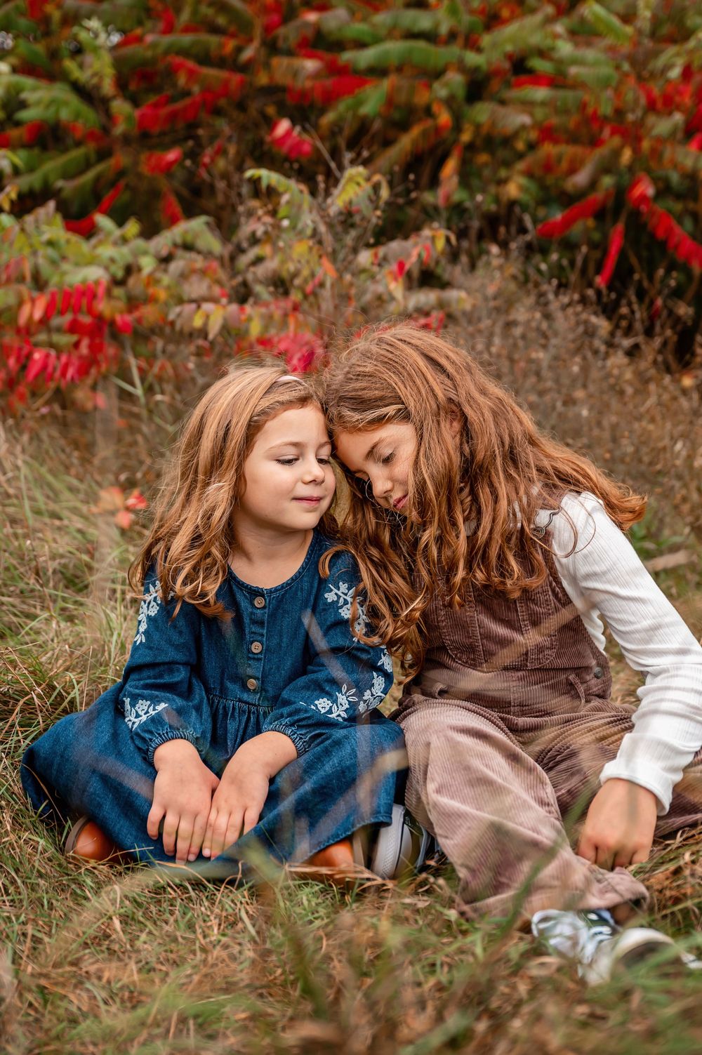 Two young friends in casual clothing sitting together on grass near red flowers on an autumn day.