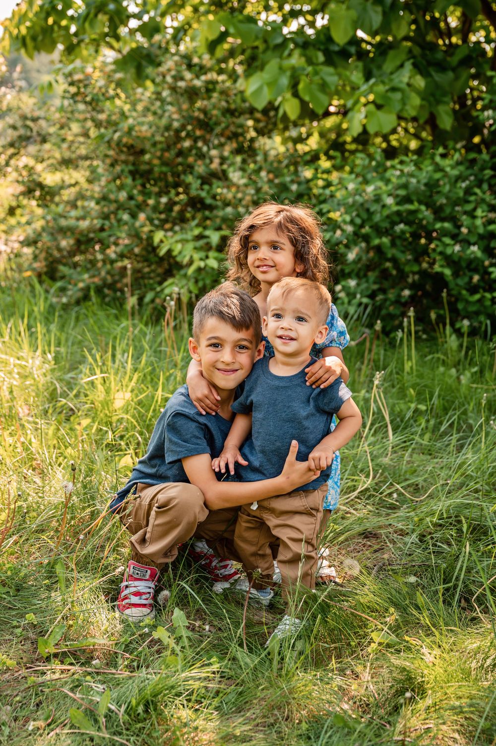 Three children in casual clothing pose together on grassy hill during golden hour in nature setting.