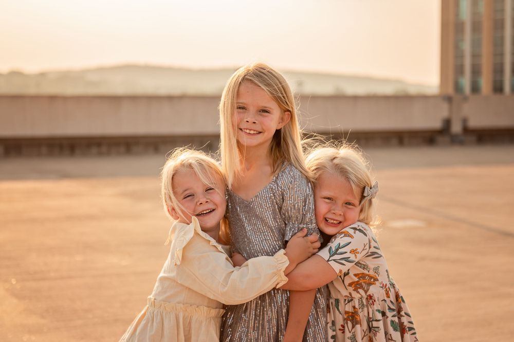 Three children embrace and smile together in warm sunset lighting.