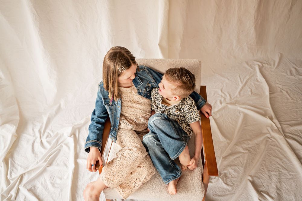 Two people share a tender moment while sitting on a white bedspread in matching denim outfits.