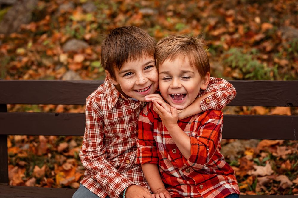 Two children in plaid shirts laughing together on a wooden bench surrounded by autumn leaves.