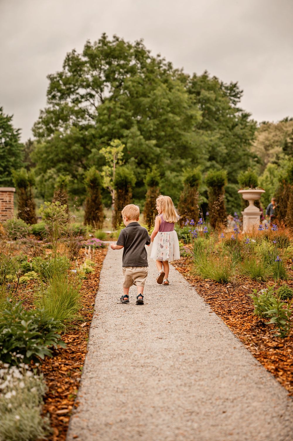 Three people walk down a gravel garden path lined with autumn leaves and colorful perennial plantings.