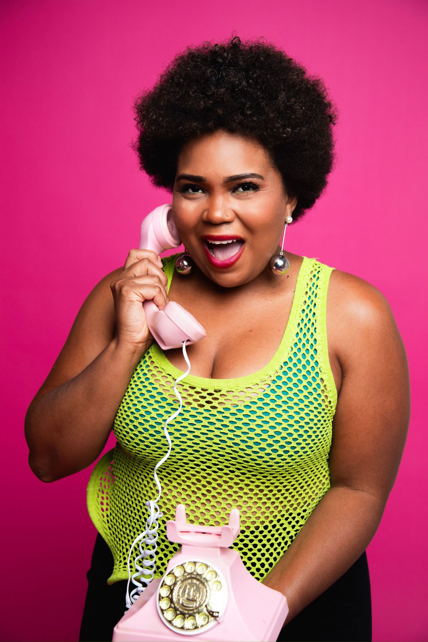 Woman in neon mesh top holding pink retro telephone against bright pink background.