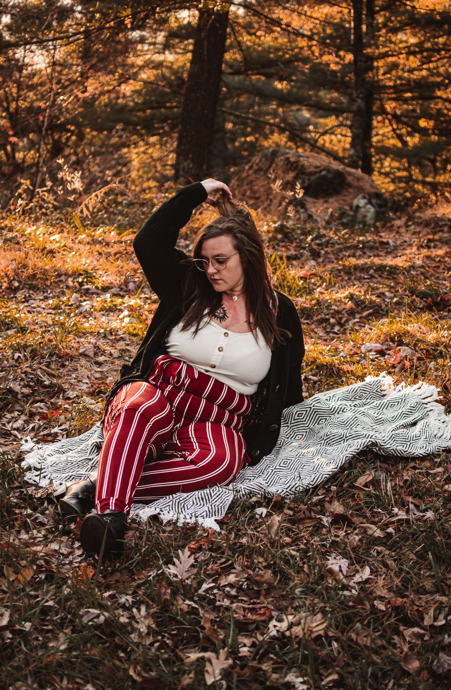 Person in striped pants sits on blanket amid fallen leaves in golden-lit autumn forest.