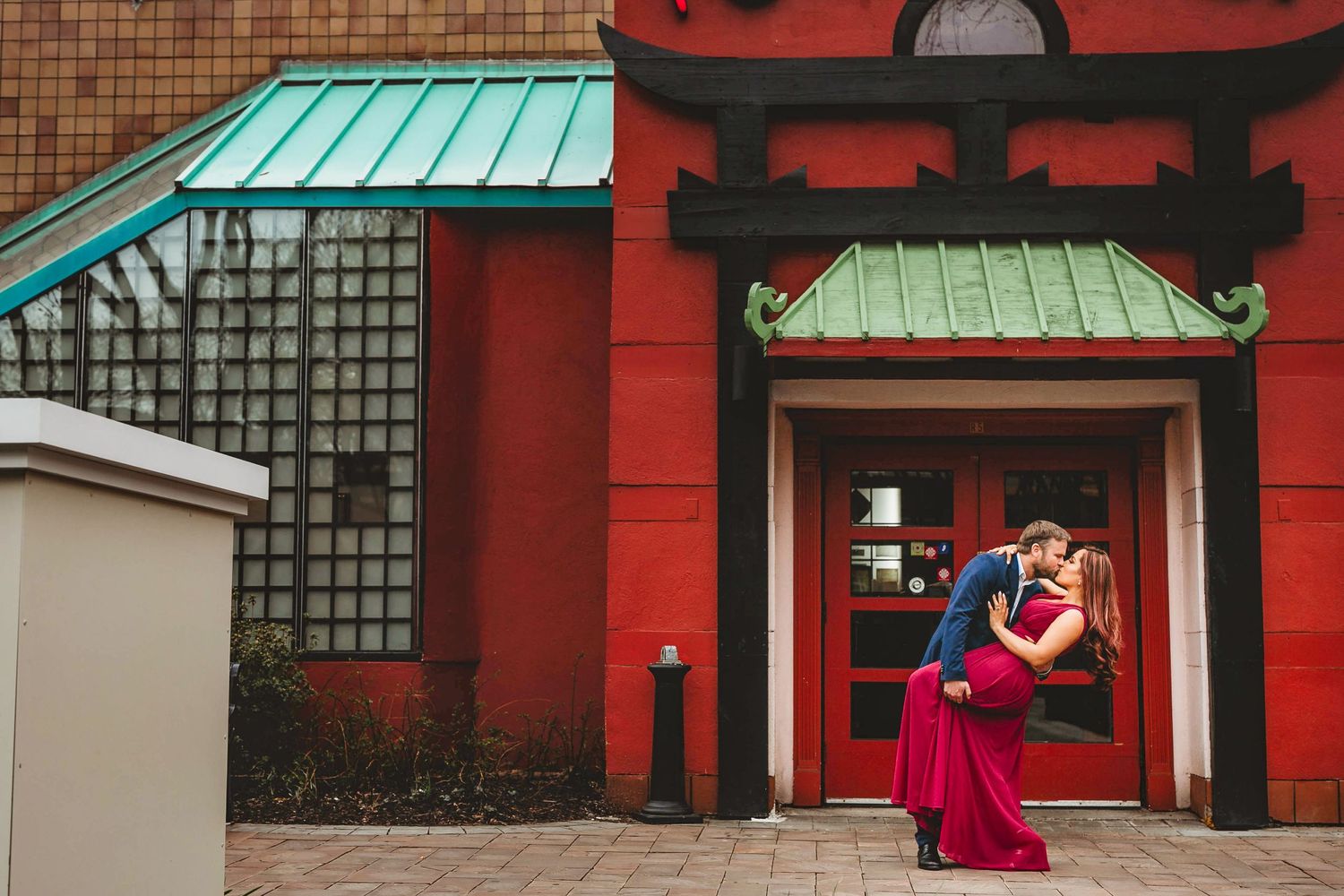 Couple sharing a romantic dip kiss in front of a red Asian-inspired building during an engagement photo shoot.