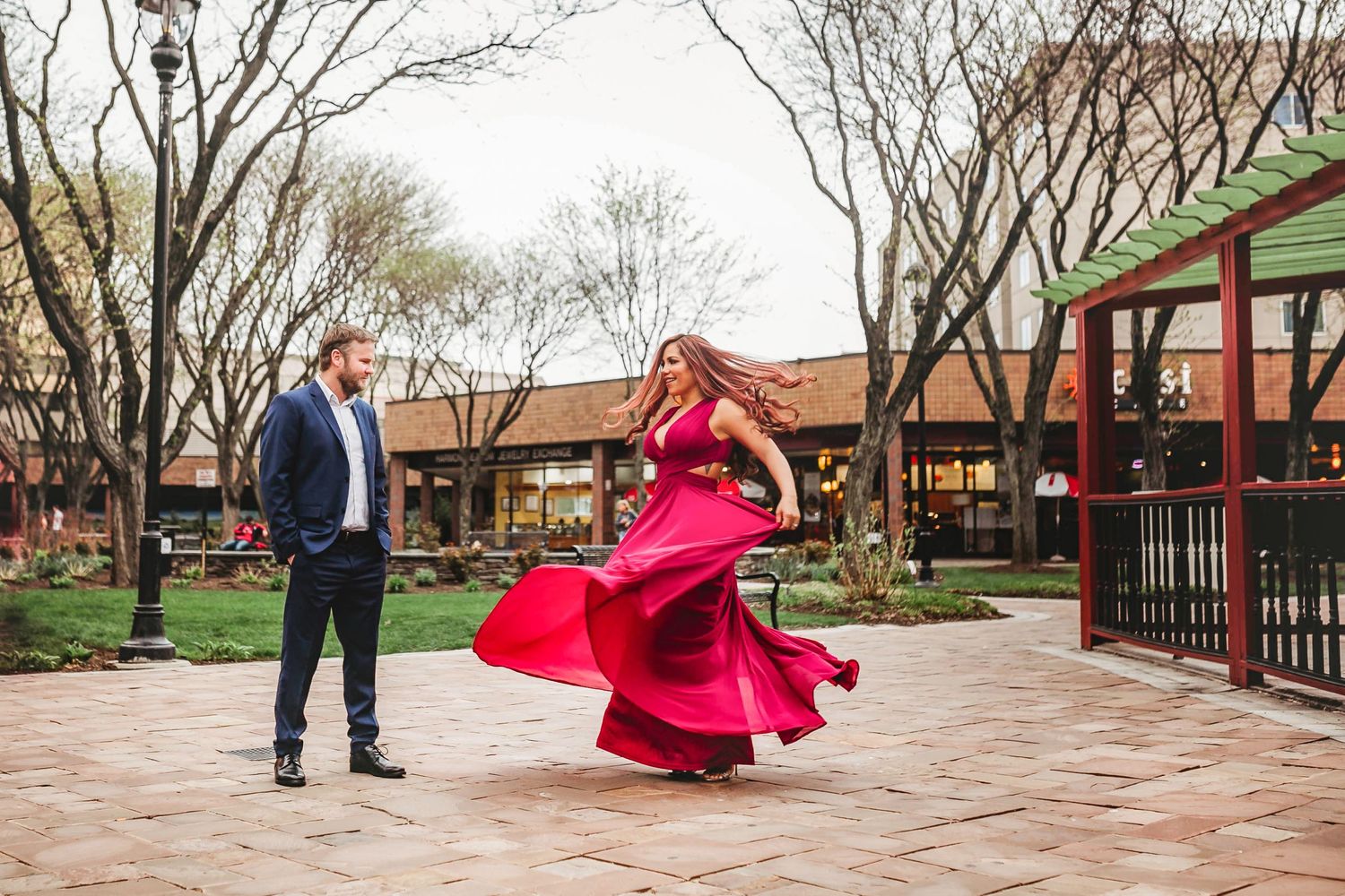 Woman in a flowing red gown twirling while her partner watches during a vibrant outdoor engagement photo session.