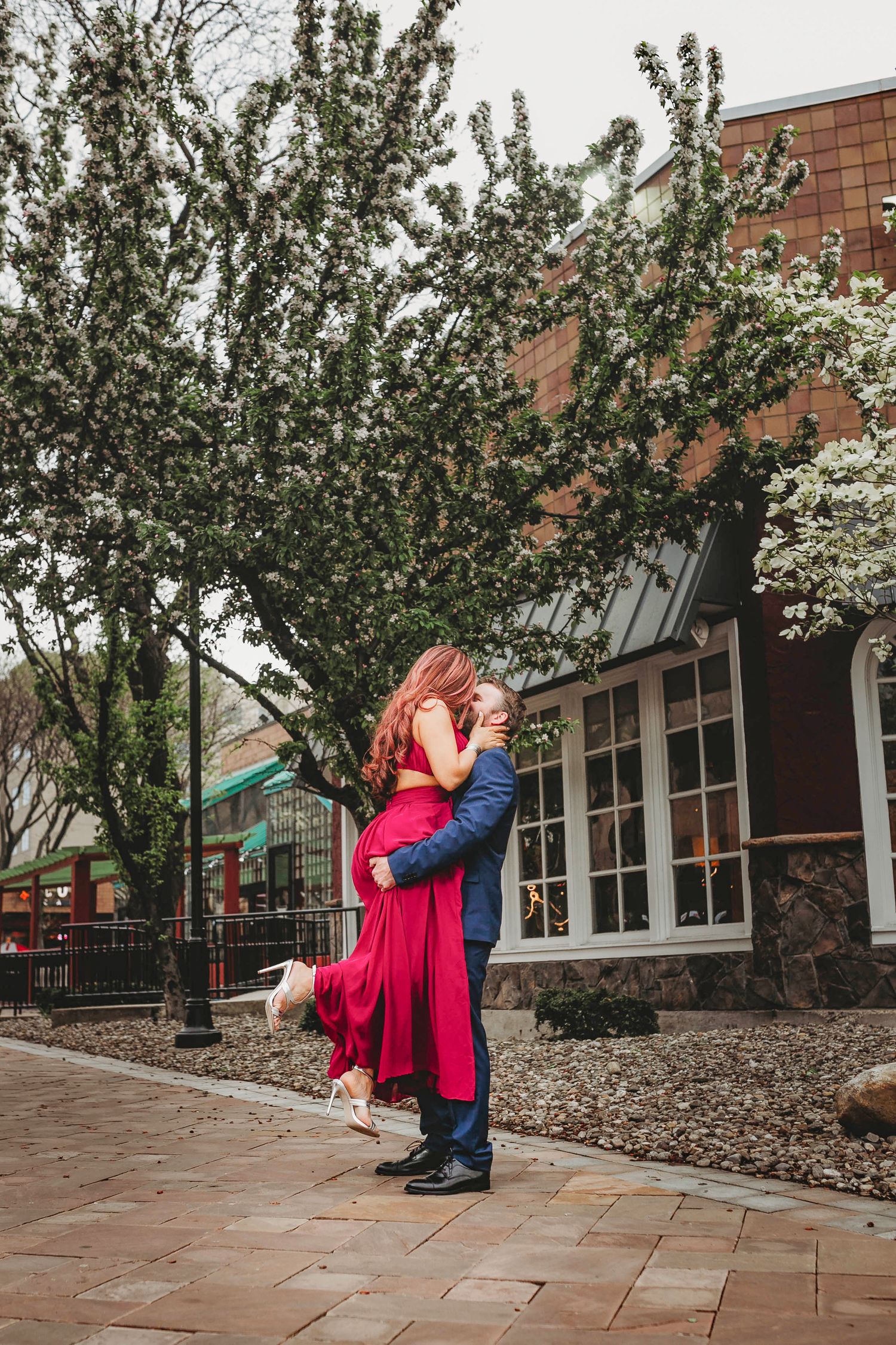 Groom lifts bride in red dress for a kiss under blooming trees in outdoor engagement photo.