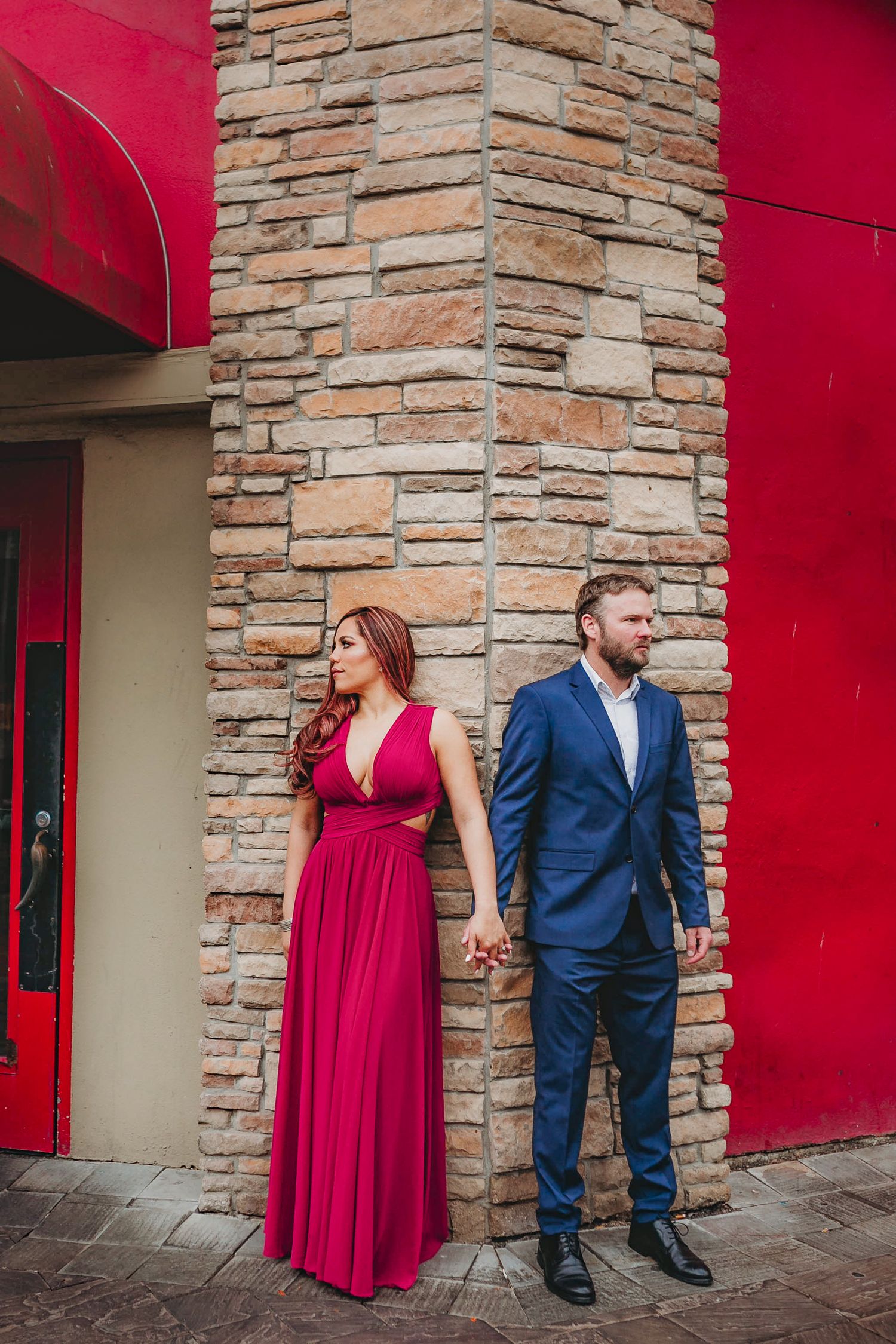 Engaged couple holding hands by stone wall, woman in red gown and man in blue suit.