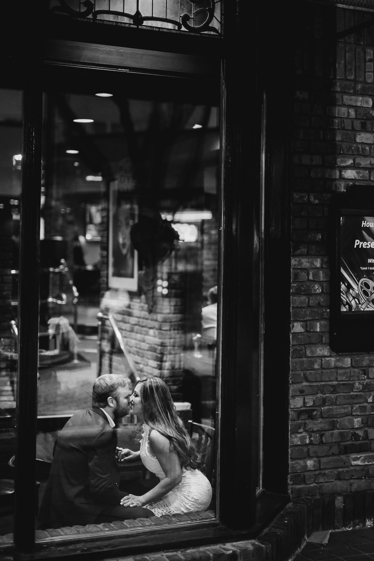 Romantic couple kissing inside a cozy brick restaurant viewed through a window at night.