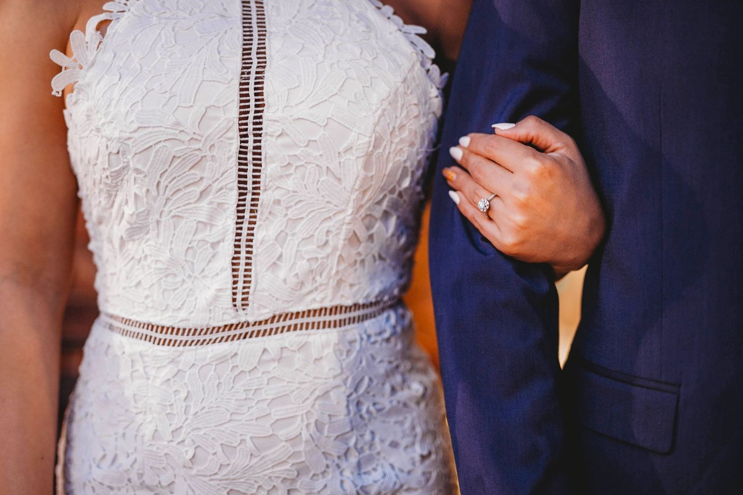 Bride in white lace halter dress with gold belt detail holds groom's arm showing engagement ring.