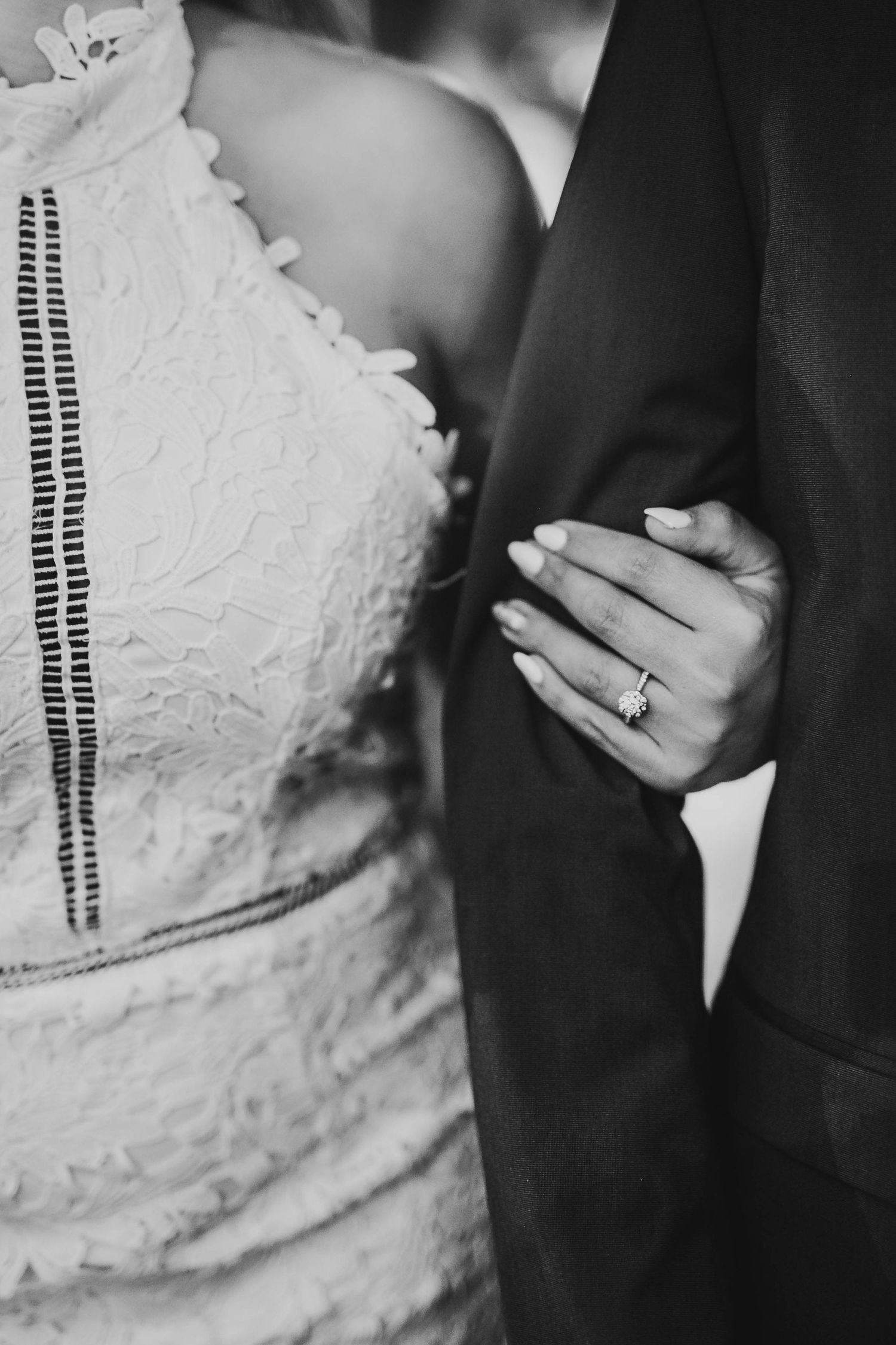 Close-up of bride in lace dress holding groom's arm showing diamond engagement ring in black and white.