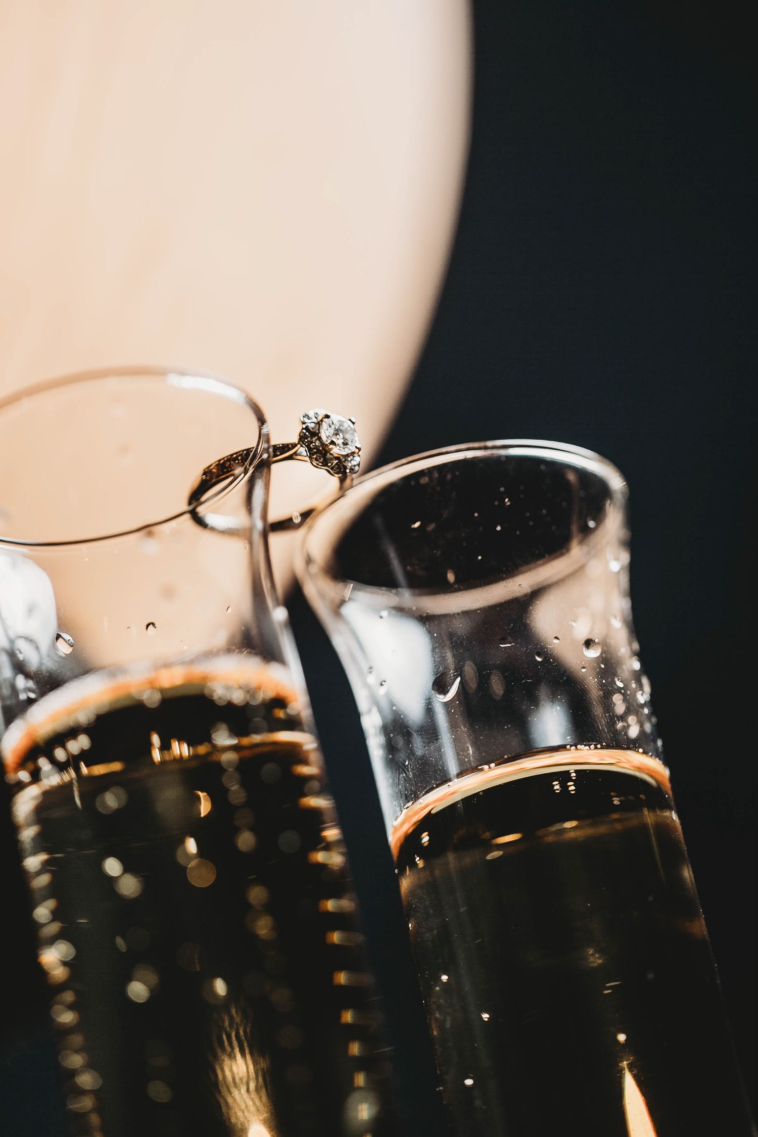 Diamond engagement ring balanced on champagne flutes filled with sparkling wine on dark background.