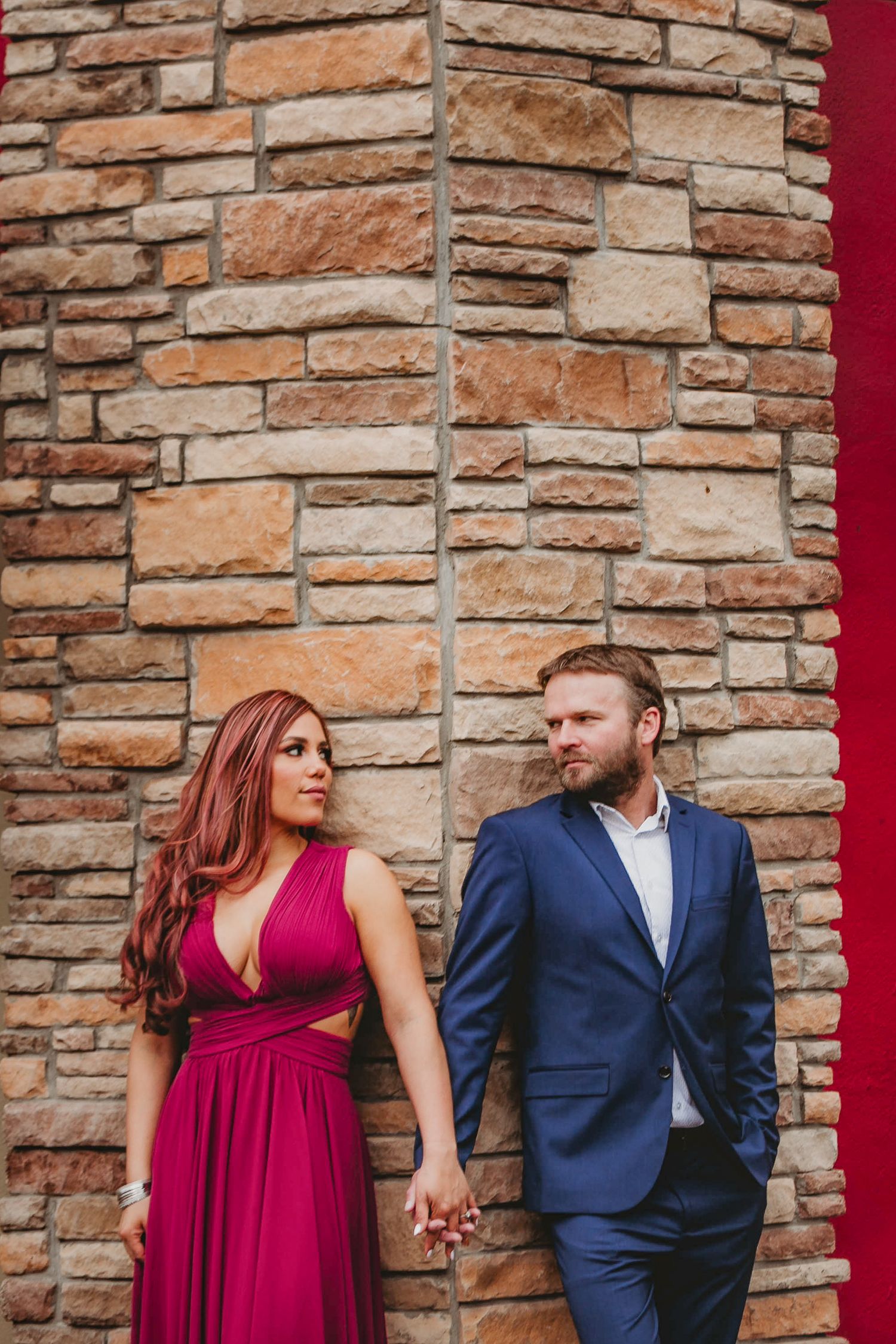 Couple holding hands against rustic stone wall, woman in magenta dress and man in navy suit.
