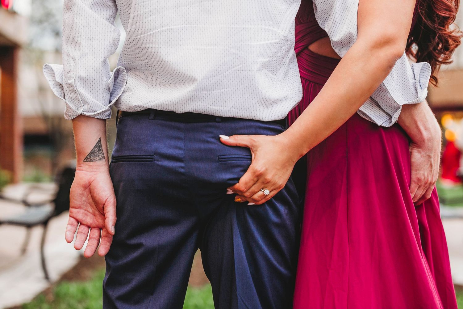 Woman in red dress slips hand into man's suit pocket during outdoor engagement photo session.