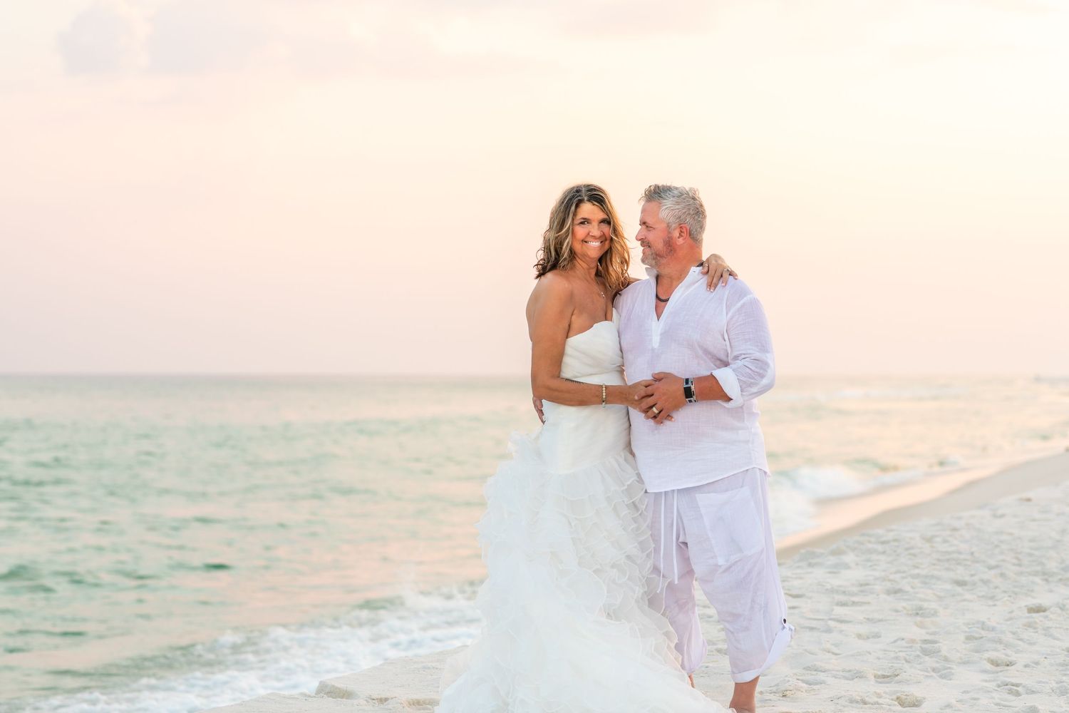 A romantic beach wedding photoshoot at sunset with couple walking along the shore in white attire.