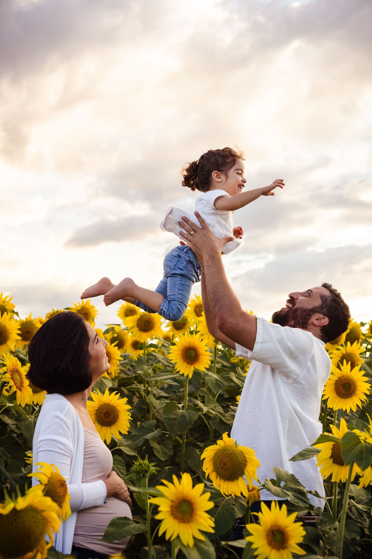 family on a sunflower field dad is holding his daughter in the air and the pregnant mom is smiling at them