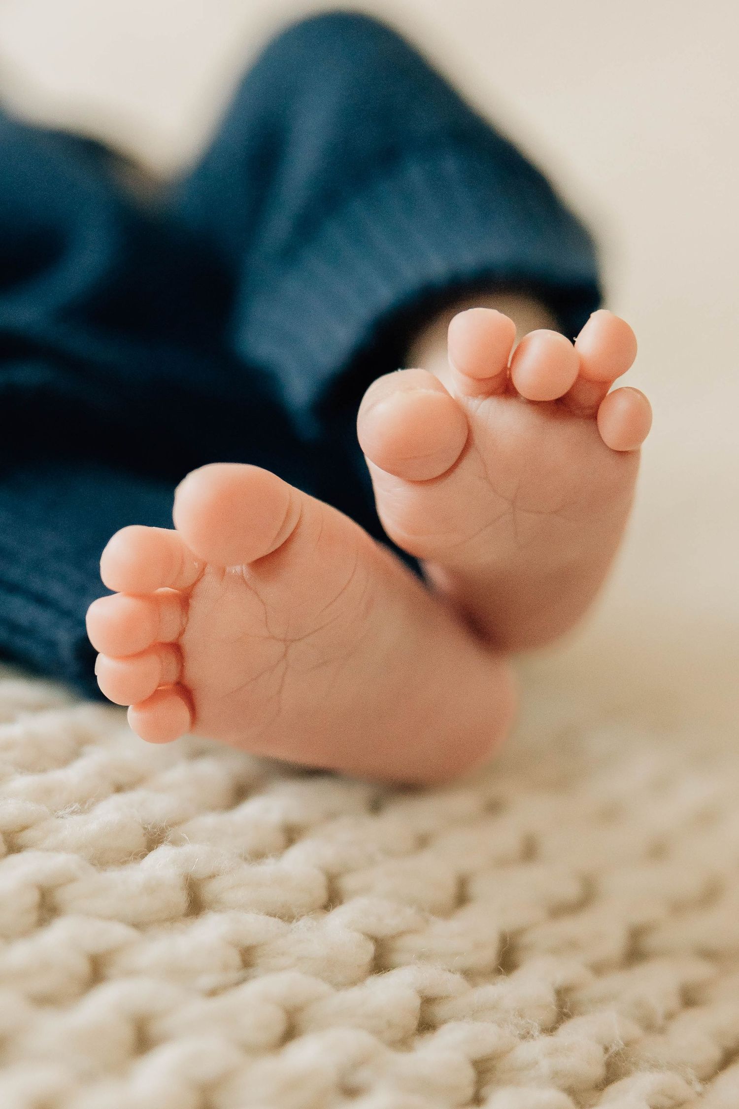 Close-up of tiny newborn baby feet in a blue knit outfit resting on a cream textured blanket.