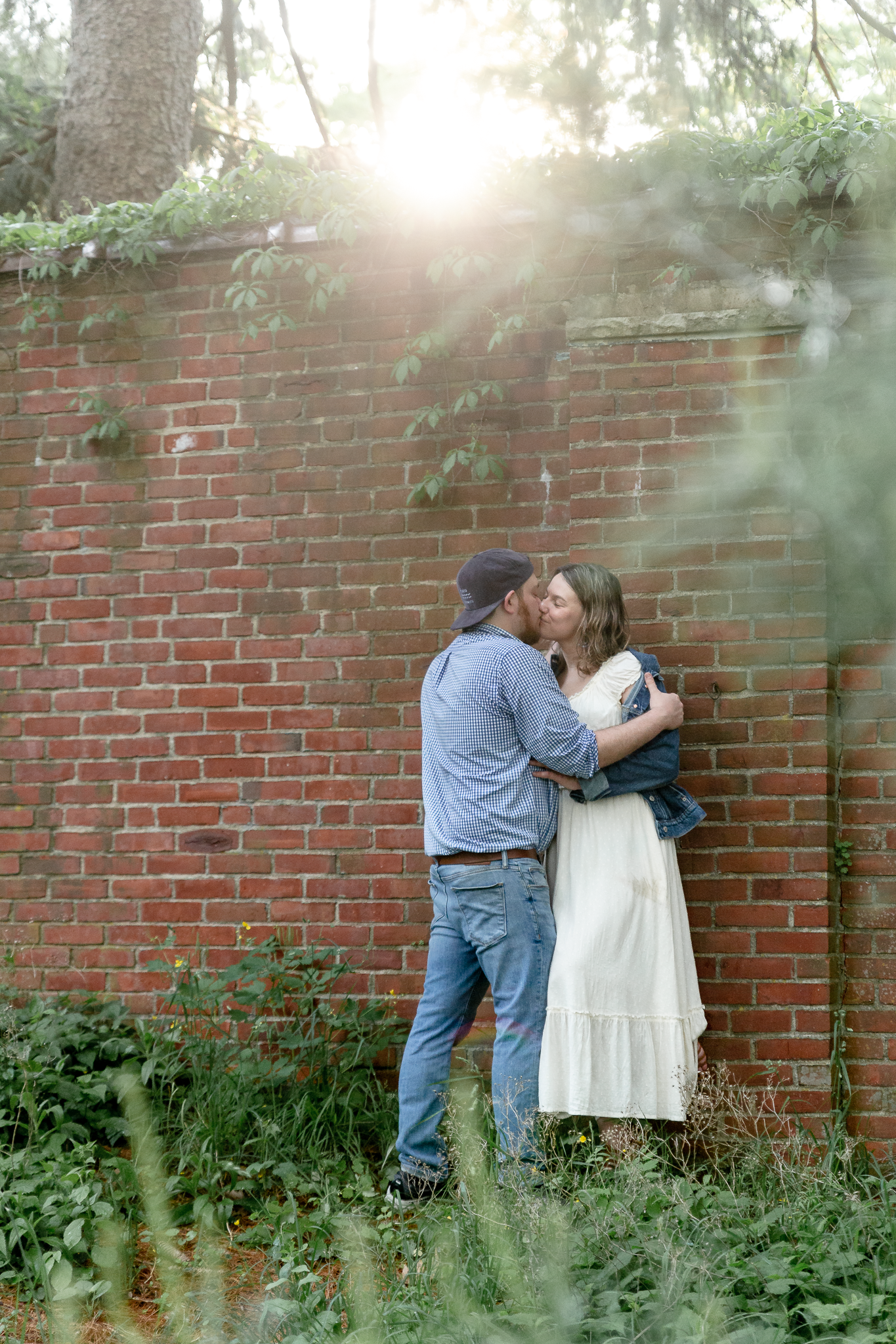 woman in white dress and denim jacket kissing man in blue shirt below the sun against ivy covered brick wall