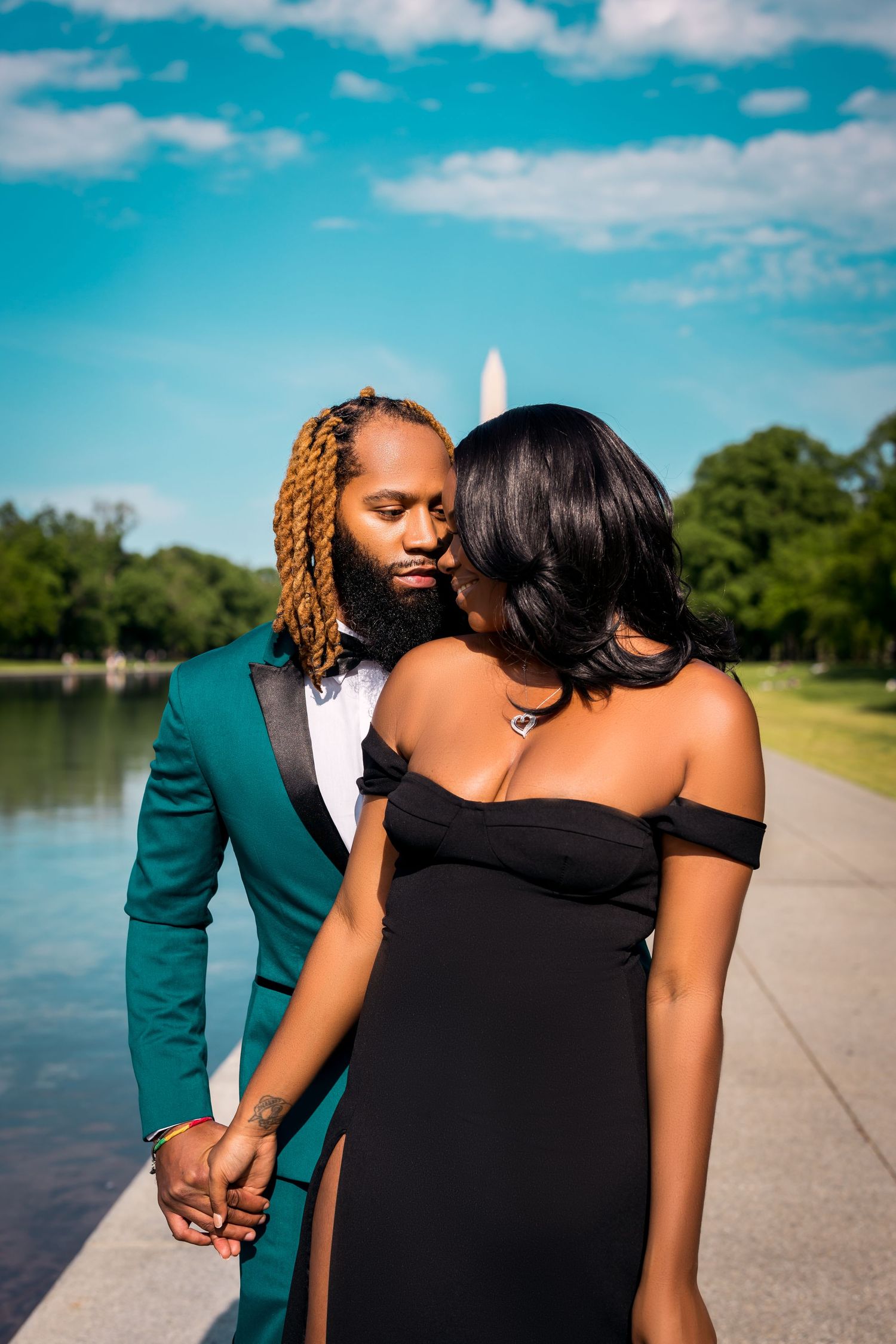 Romantic couple in formal attire poses near reflecting pool with trees in background.