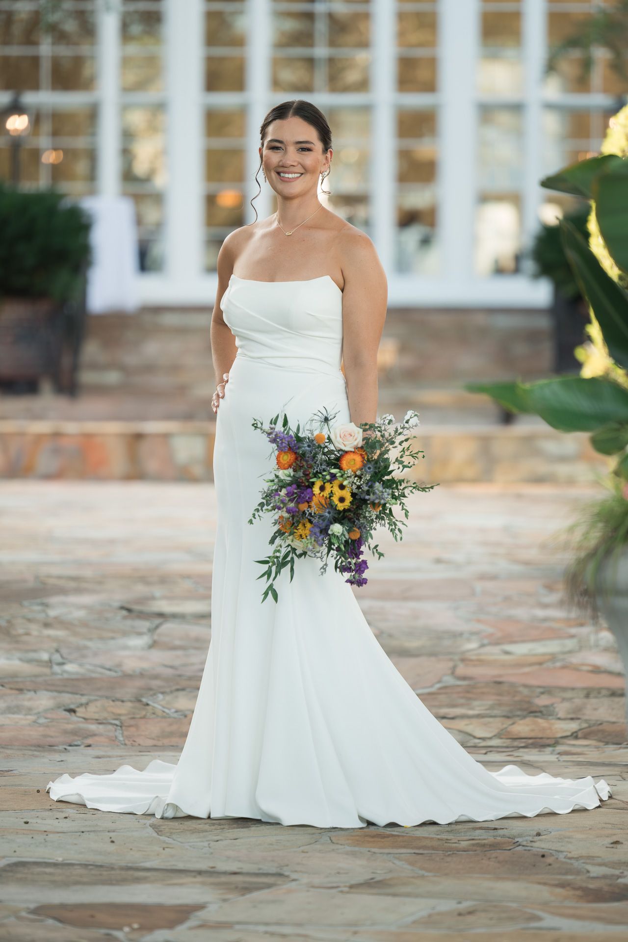 Bride in elegant white gown holds vibrant wildflower bouquet on stone patio.