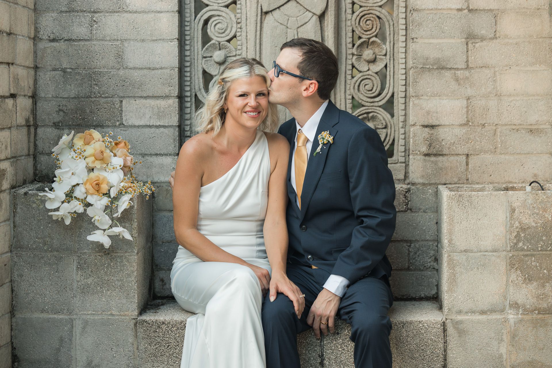 Newlyweds share intimate moment against ornate stone wall.