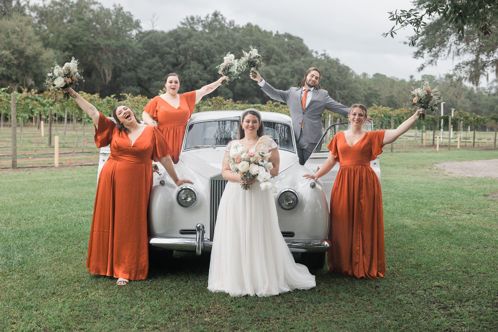 Bridesmaids in burnt orange dresses posing playfully with bride near vintage car.