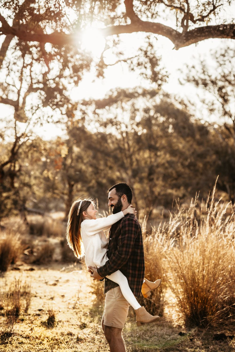 Couple embracing in romantic pose during golden hour beneath sunlit tree branches.